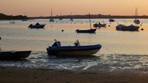 Christchurch boats This image shows a landscape view of Christchurch in Dorset, United Kingdom, taken during an evening in late spring. The main subject is transport by boats, with various vessels resting in shallow water and on the mudflats as the tide is out. The scene captures several types of boats, including small sailboats and motorboats, anchored or moored in the tranquil waters. Buildings from Christchurch are faintly visible on the horizon, and the foliage on the shoreline adds context to the location. The soft evening light casts warm tones across the water and boats, highlighting the peaceful atmosphere typical of this coastal destination in Dorset.